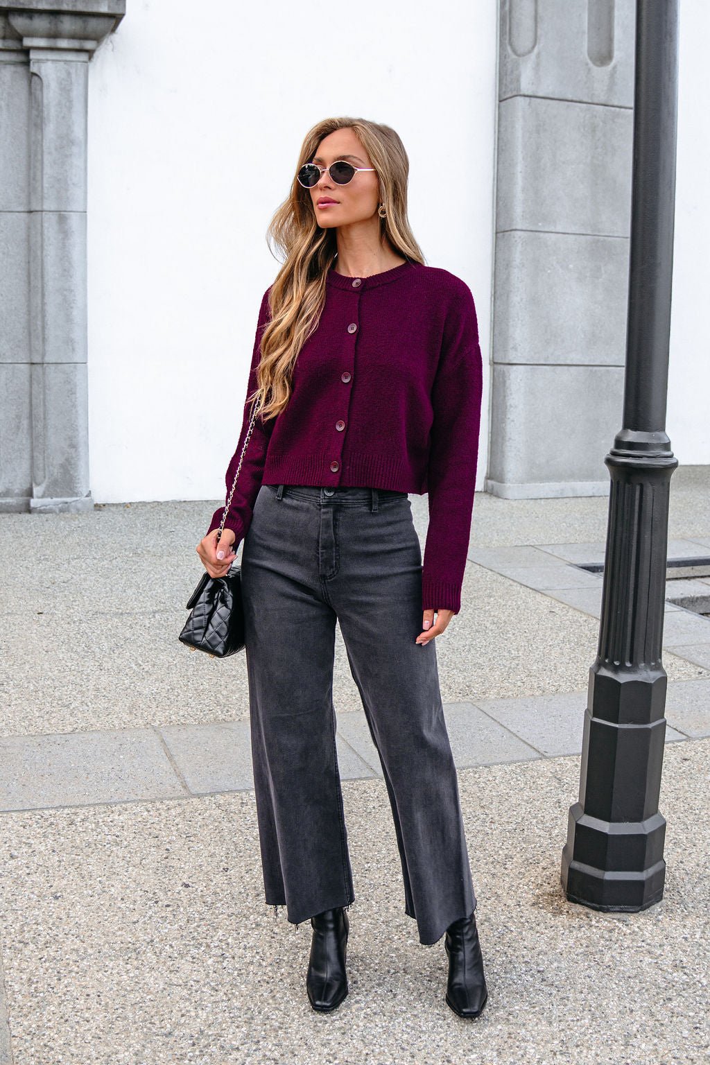 Woman in a Burgundy Ribbed Button Down Cardigan, gray jeans, and black boots stands near a lamp post with a black bag.