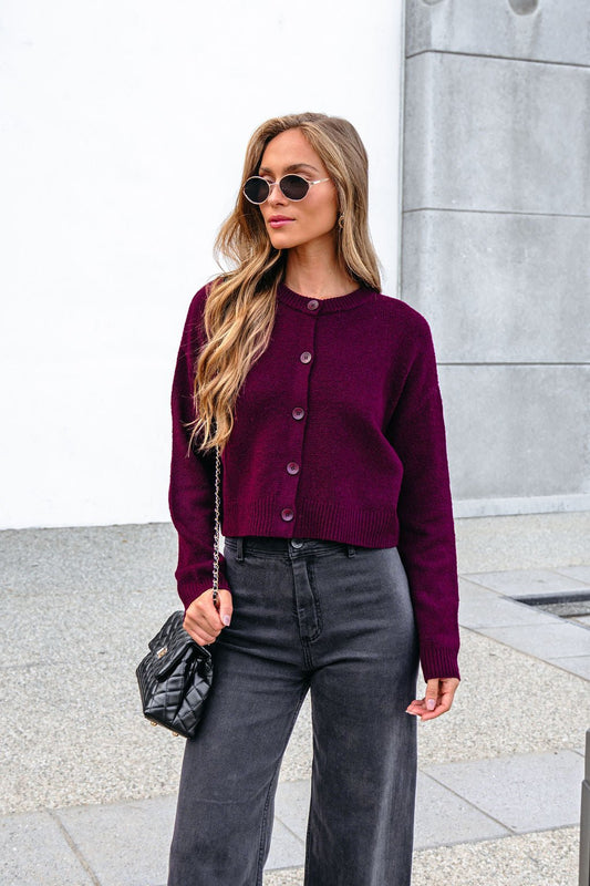 Woman in sunglasses wears a Burgundy Ribbed Button Down Cardigan, black wide-leg pants, and carries a black purse by a gray wall.