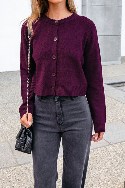 Woman in a burgundy ribbed button-down cardigan, dark gray high-waisted jeans, holding a black quilted purse.