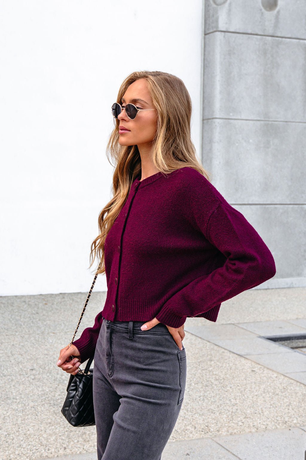 Woman in sunglasses wears a burgundy ribbed button down cardigan and gray jeans, holding a black purse by a modern wall.