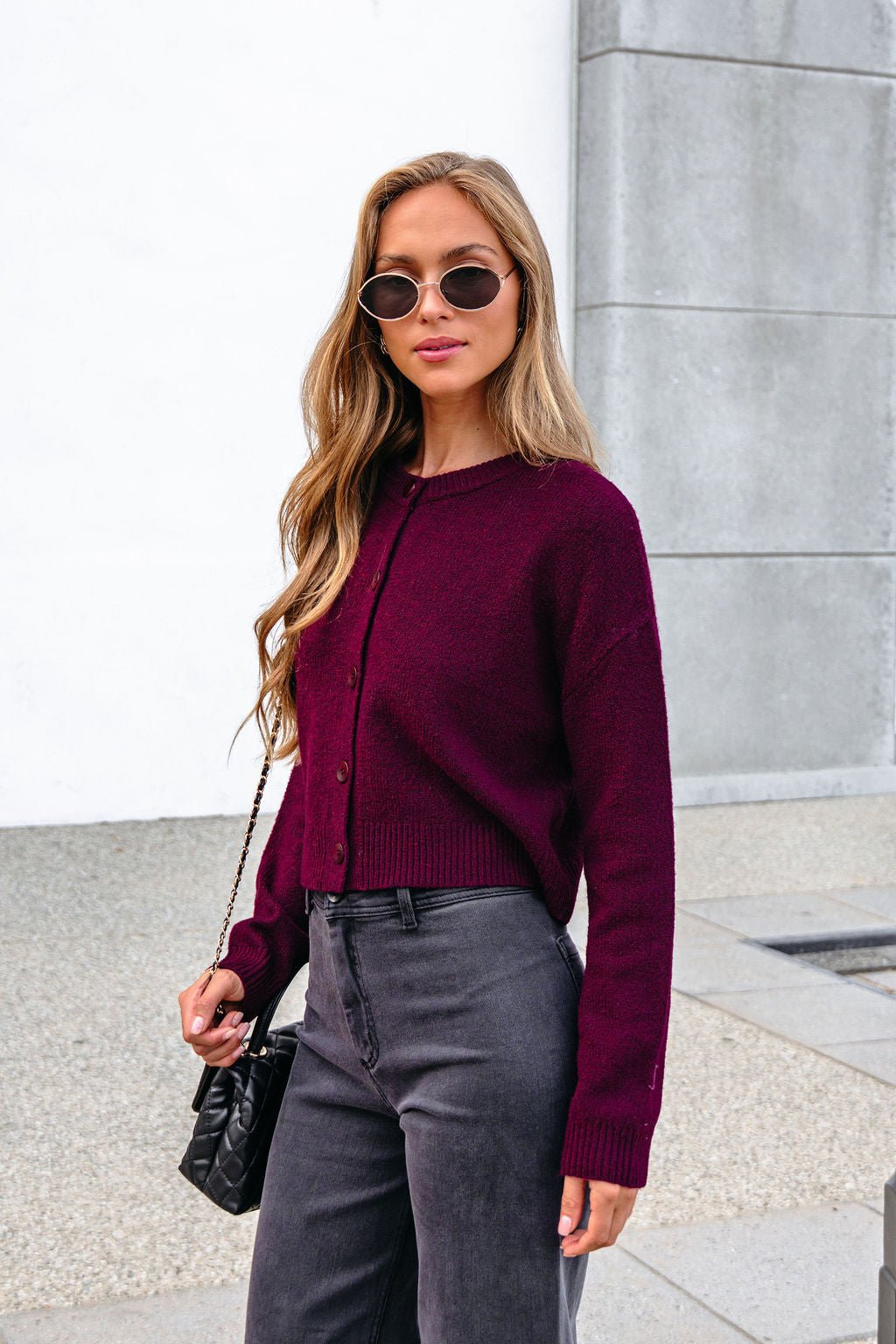 Woman with long hair in sunglasses wears a Burgundy Ribbed Button Down Cardigan and gray pants by a gray concrete wall outdoors.