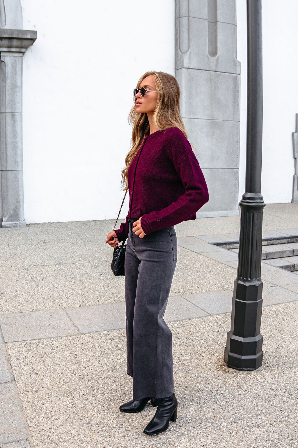Woman wearing sunglasses, Burgundy Ribbed Button Down Cardigan, gray pants, and black boots stands by a streetlamp on a stone sidewalk.