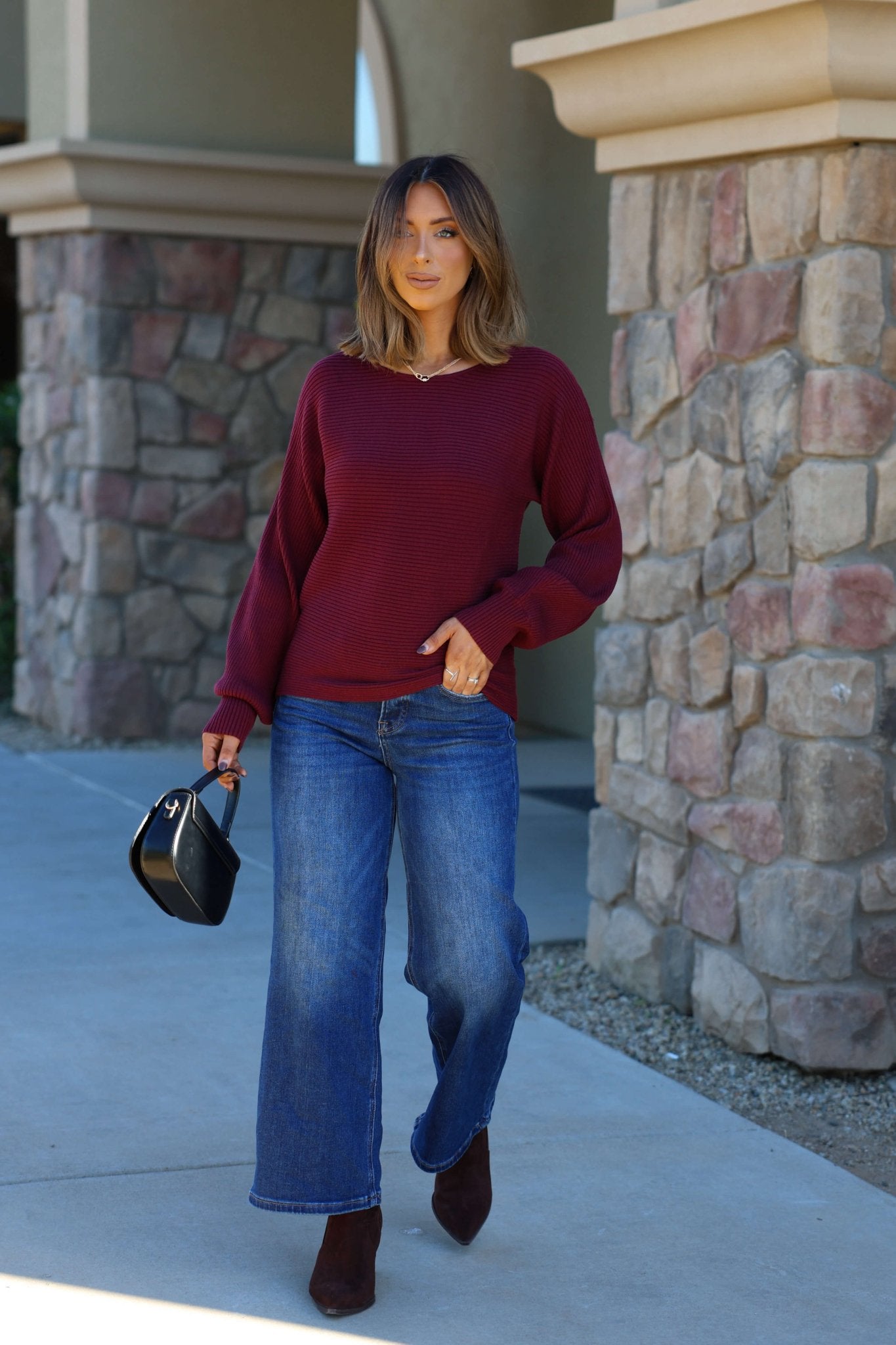 Woman with shoulder-length hair in a Burgundy Ribbed Dolman Sleeve Sweater, blue jeans, ankle boots, and a small black bag outdoors.