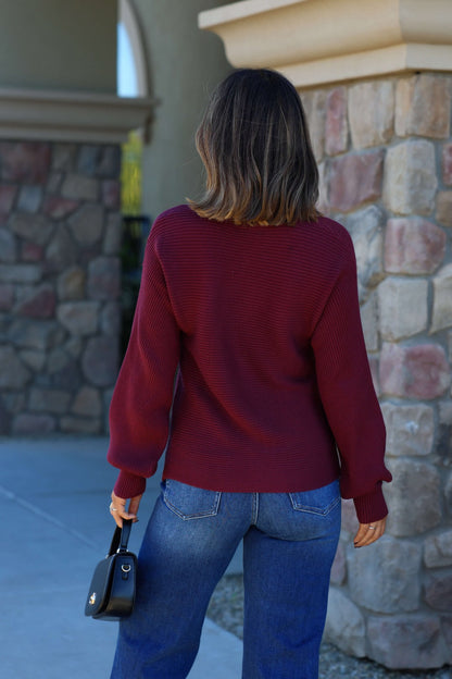 A woman in a Burgundy Ribbed Dolman Sleeve Sweater and blue jeans stands outdoors, facing away, holding a small black handbag.