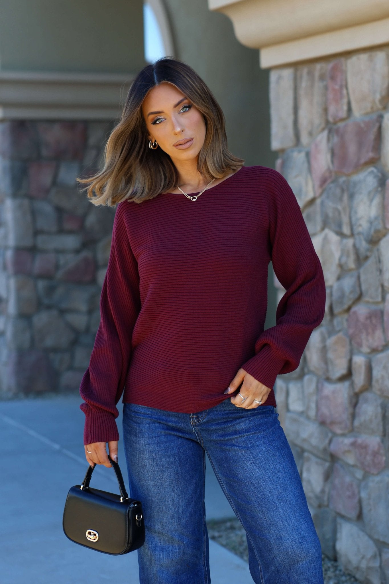 Woman in a Burgundy Ribbed Dolman Sleeve Sweater and blue jeans stands by a stone wall, holding a black handbag.