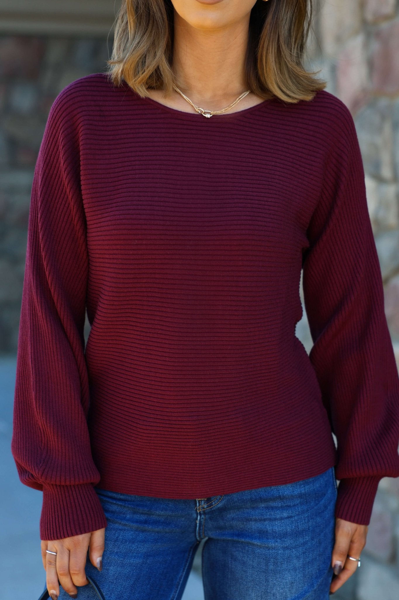 A woman stands outdoors with her hands at her sides, wearing a Burgundy Ribbed Dolman Sleeve Sweater and blue jeans.