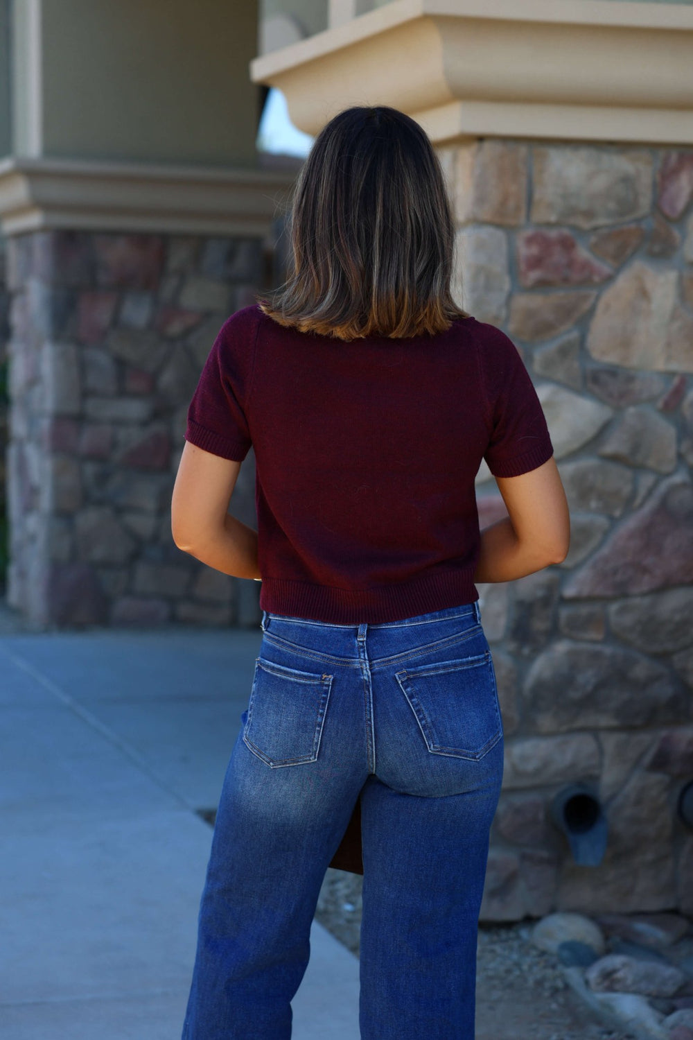 A woman in a Burgundy Short Sleeve Ribbed Sweater - FINAL SALE and blue jeans stands outside by a stone building.