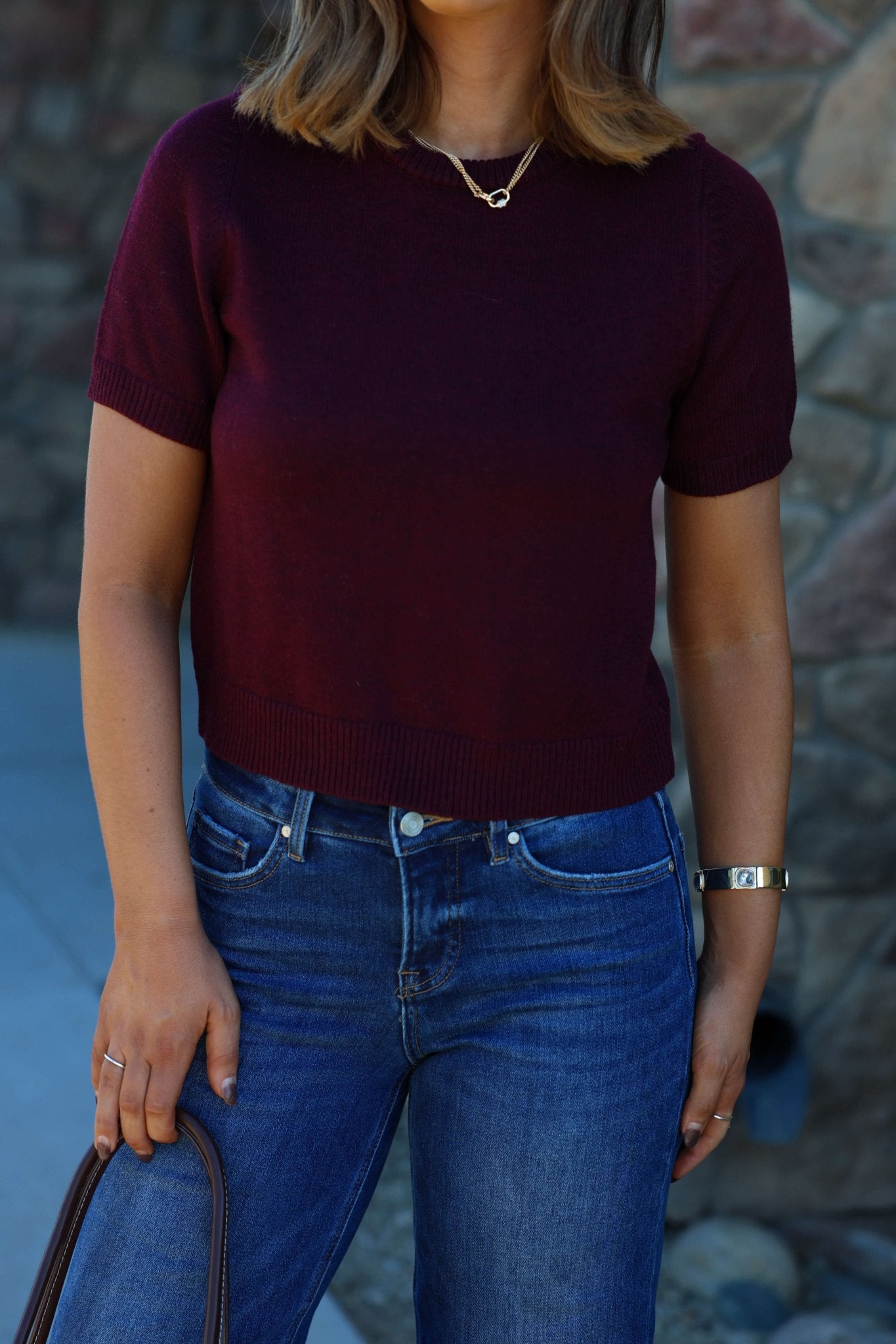 A woman in a Burgundy Short Sleeve Ribbed Sweater and blue jeans holds a brown handbag by a stone wall.