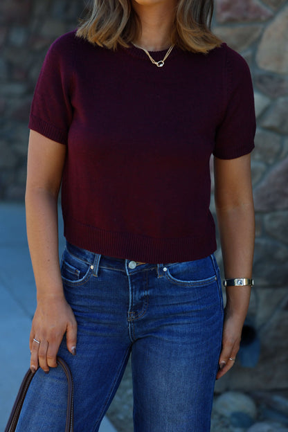 A woman in a Burgundy Short Sleeve Ribbed Sweater and blue jeans holds a brown handbag by a stone wall.