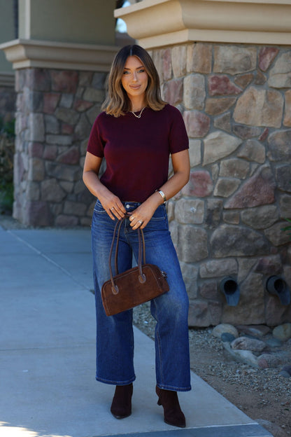 Woman in front of a stone wall wearing a Burgundy Short Sleeve Ribbed Sweater, blue jeans, and holding a brown handbag.