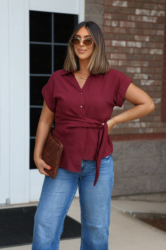 Woman in sunglasses wears a Burgundy Side Tie Buttoned Top and jeans, holding a brown clutch near a brick wall.