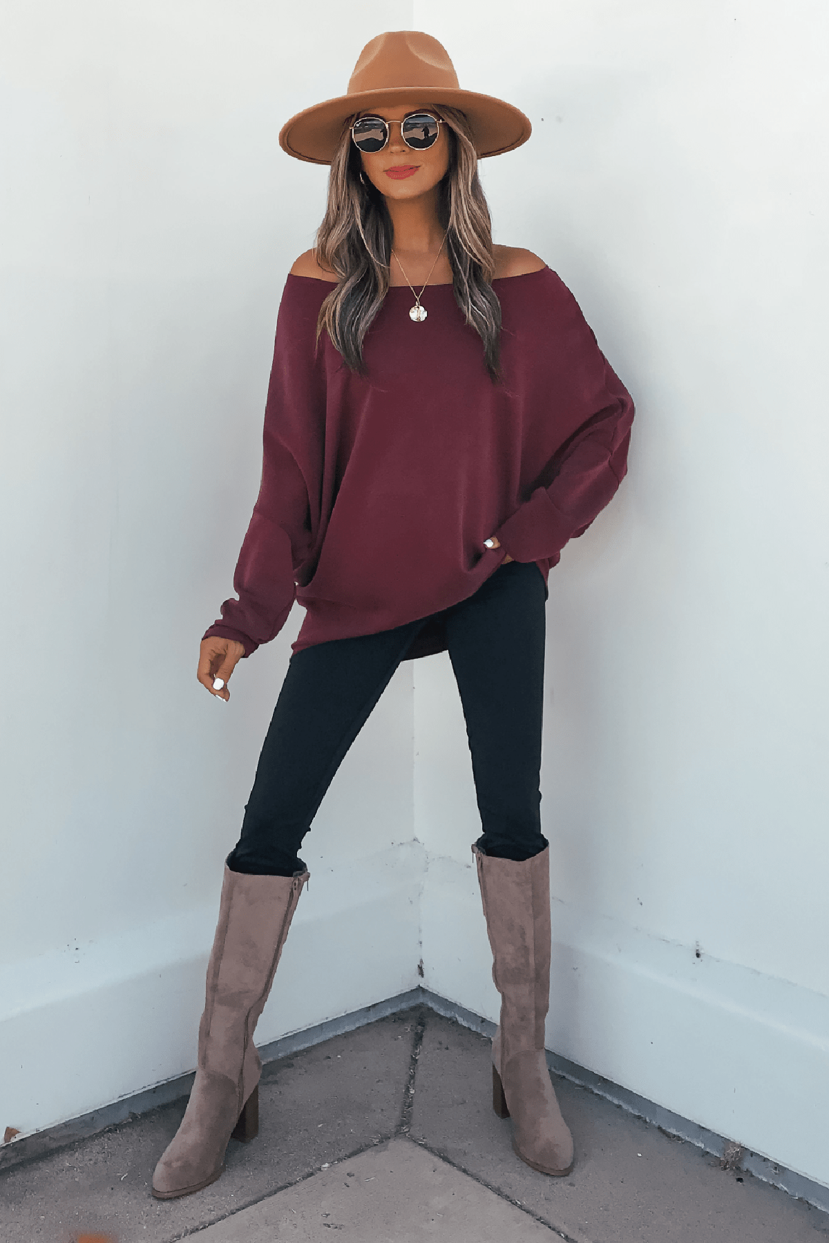 Woman in a tan hat, sunglasses, and Burgundy Soft Cloud Scuba Pullover stands by a white wall in black pants and taupe boots.