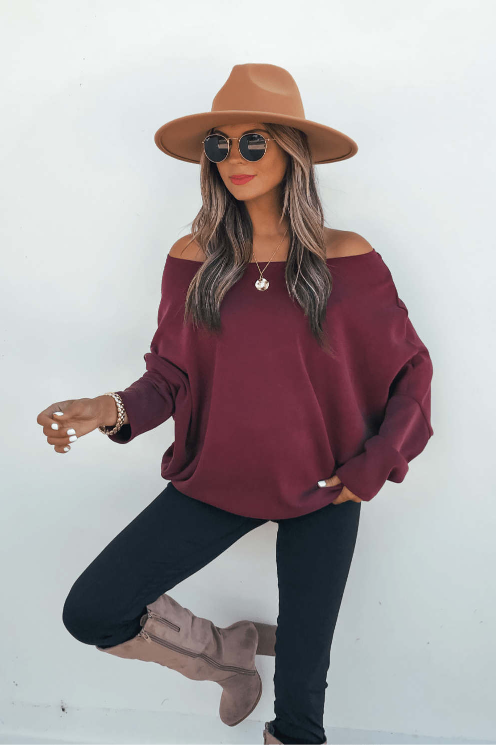 Woman in sunglasses, Burgundy Soft Cloud Scuba Pullover, black pants, taupe boots poses stylishly against a white background.