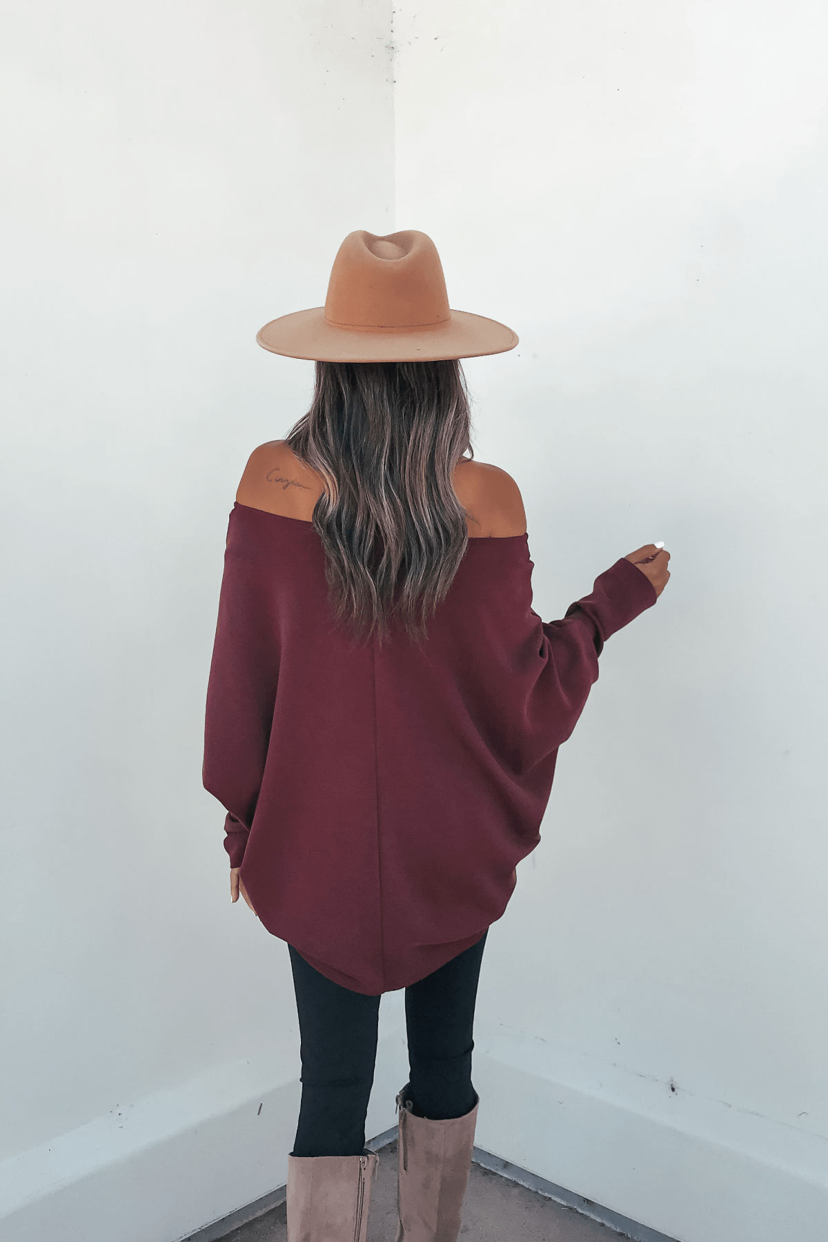 Woman with long wavy hair in a Burgundy Soft Cloud Scuba Pullover, tan hat, black pants, and tan boots facing a white wall.