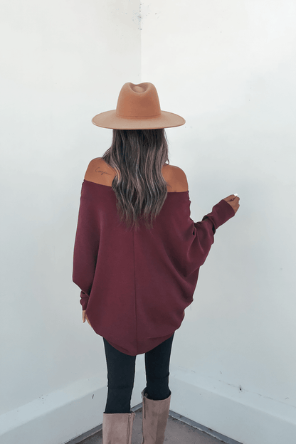 Woman with long wavy hair in a Burgundy Soft Cloud Scuba Pullover, tan hat, black pants, and tan boots facing a white wall.
