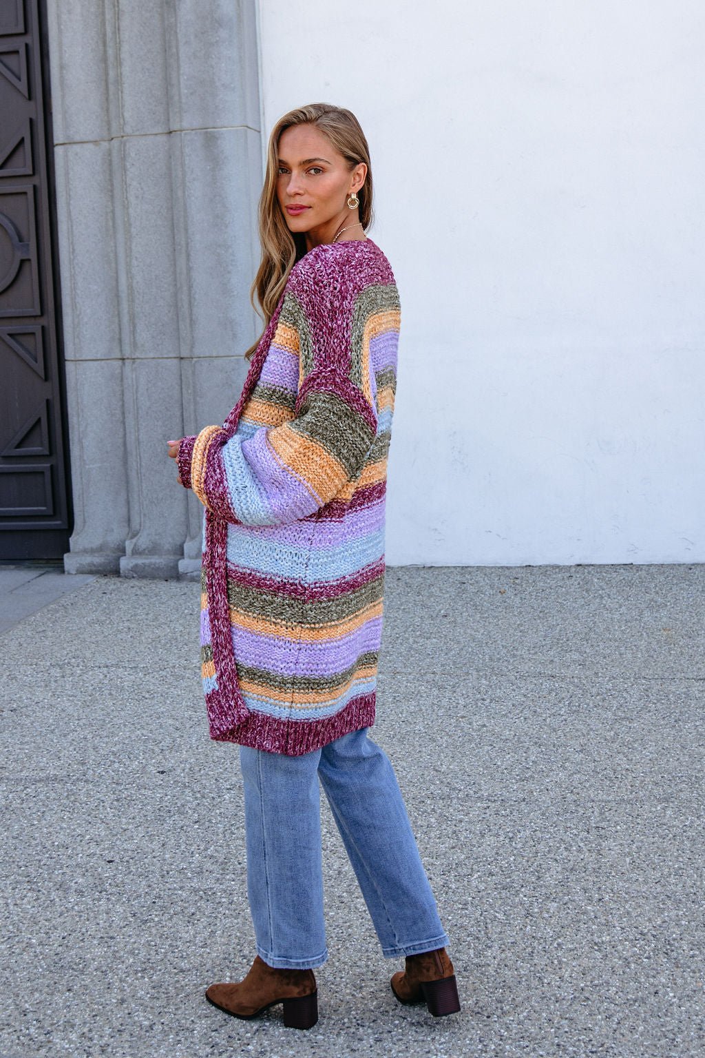 Woman in a Burgundy Striped Open Front Cardigan with jeans and boots stands by a gray stone wall, glancing back.
