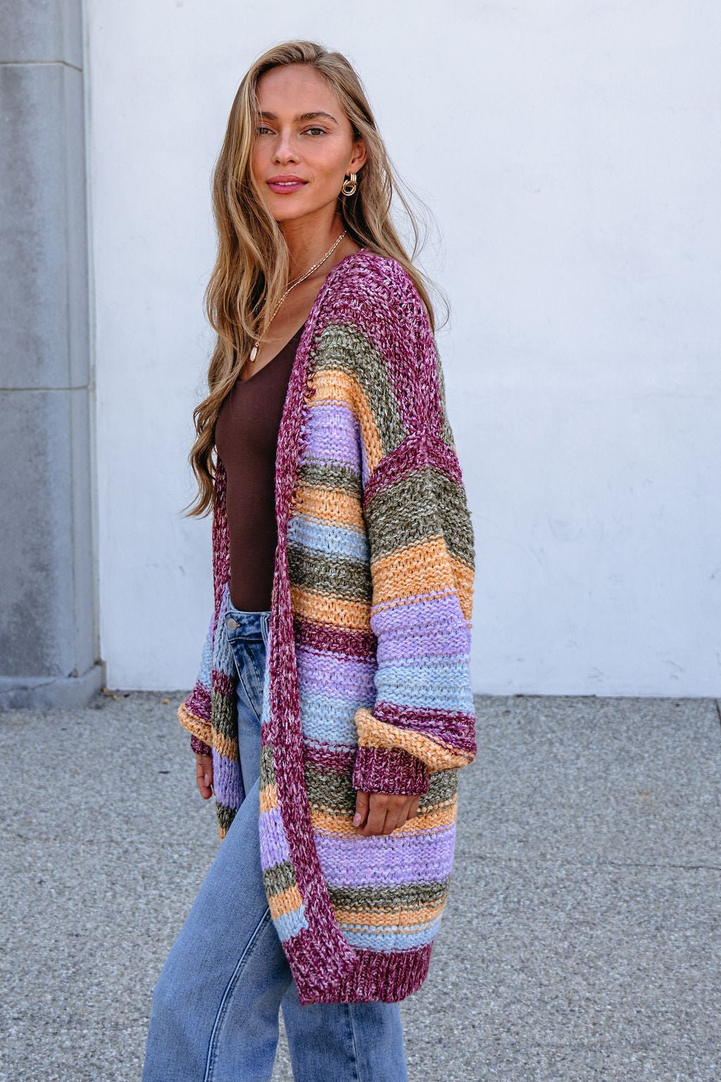 Woman in a Burgundy Striped Open Front Cardigan over a brown top and jeans, standing by a wall, looking at the camera.