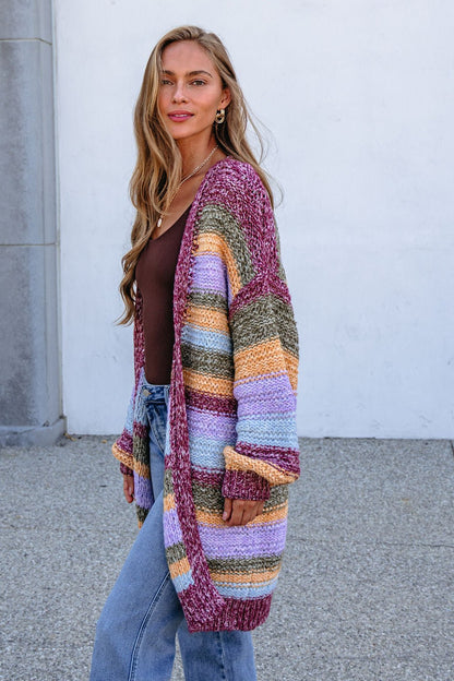 Woman in a Burgundy Striped Open Front Cardigan over a brown top and jeans, standing by a wall, looking at the camera.