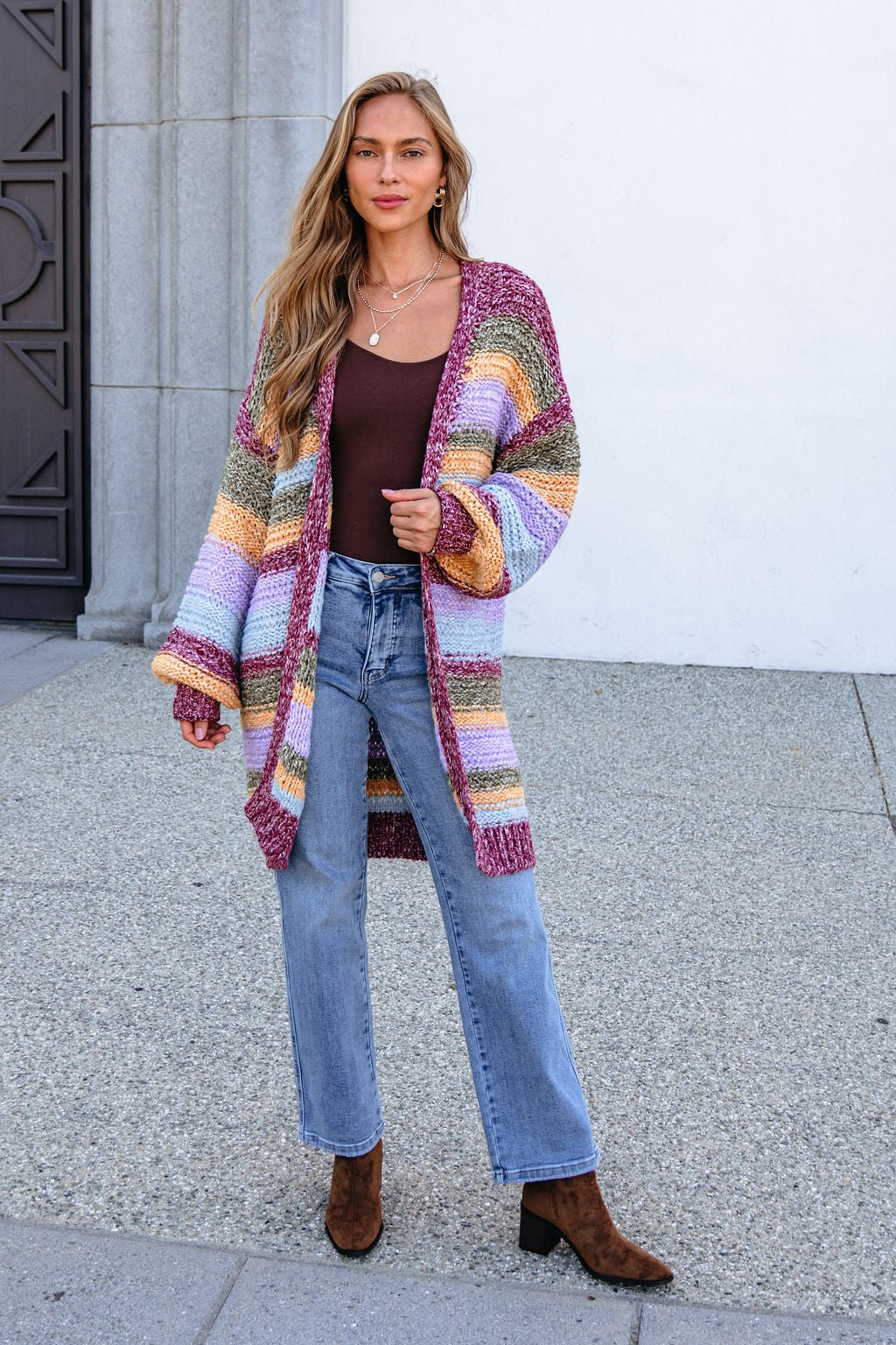 Woman in a burgundy striped open front cardigan, brown top, jeans, and boots stands by a white wall.