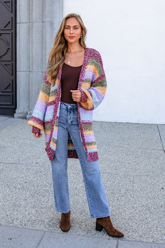 Woman in a burgundy striped open front cardigan, brown top, jeans, and boots stands by a white wall.