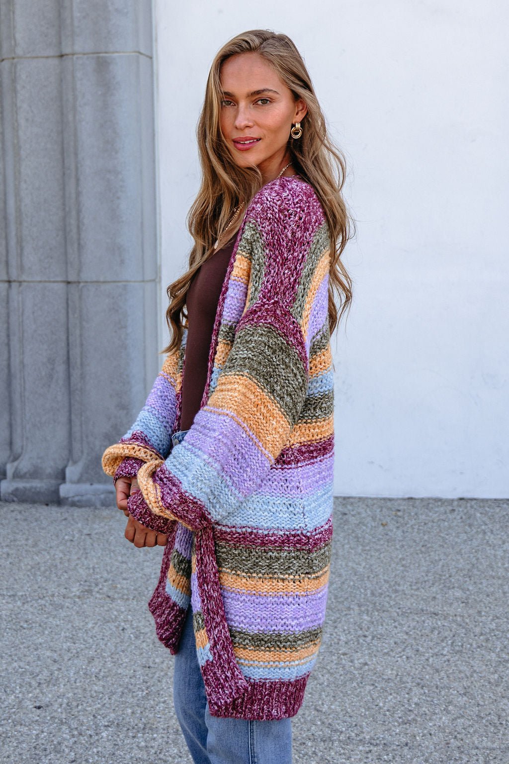 Woman with long hair in a bump-friendly Burgundy Striped Open Front Cardigan and jeans, standing by a gray stone wall.