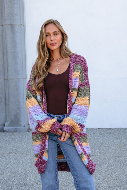 Woman in a bump-friendly Burgundy Striped Open Front Cardigan, brown top, and jeans stands outdoors by a white wall.