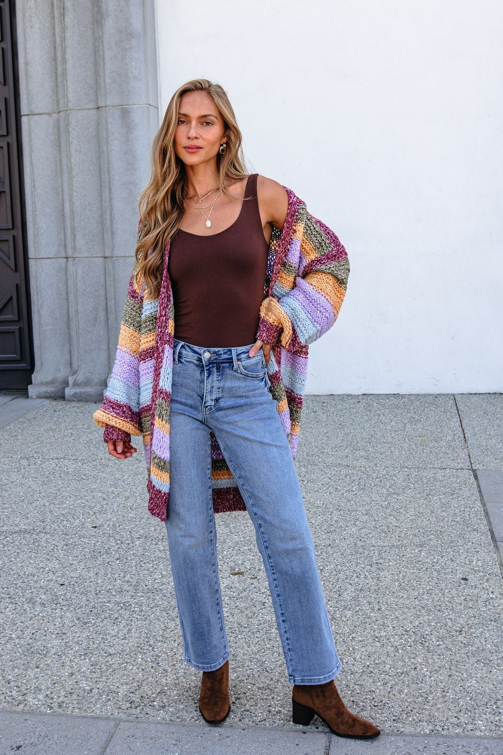 Woman in a bump-friendly burgundy striped open front cardigan, brown tank top, blue jeans, and boots, standing outdoors by a building.