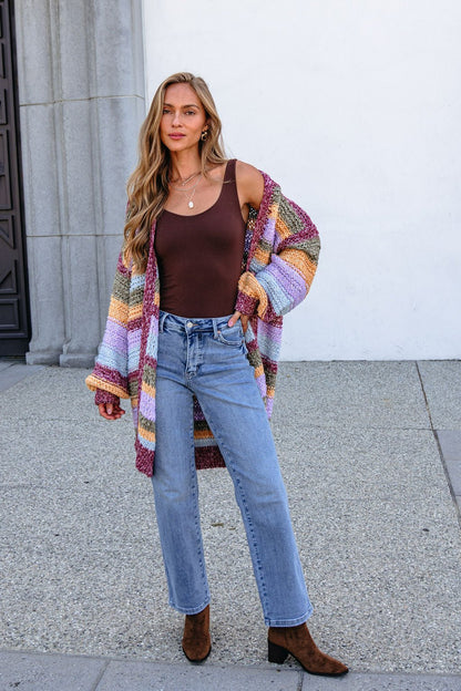 Woman in a bump-friendly burgundy striped open front cardigan, brown tank top, blue jeans, and boots, standing outdoors by a building.