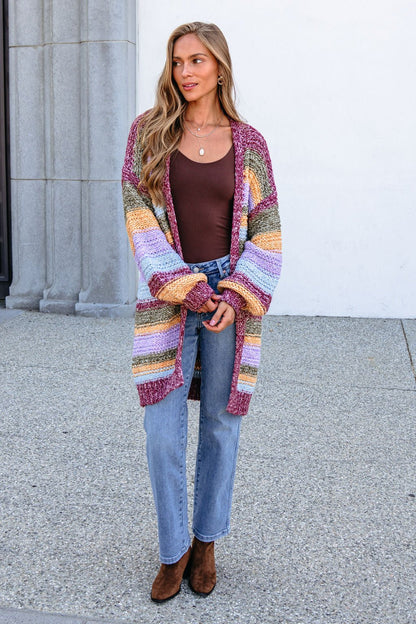 Woman in Burgundy Striped Open Front Cardigan, brown top, and blue jeans stands on the sidewalk with a cozy sweater knit look.