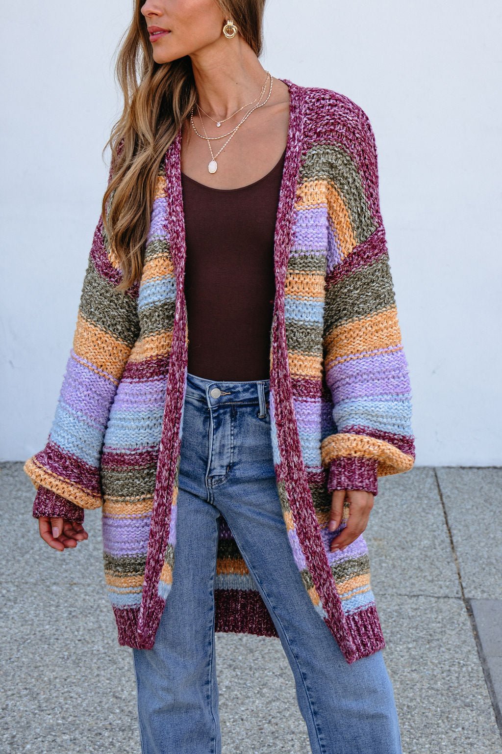 Woman in a bump-friendly Burgundy Striped Open Front Cardigan, brown top, blue jeans, and gold jewelry, standing outdoors.