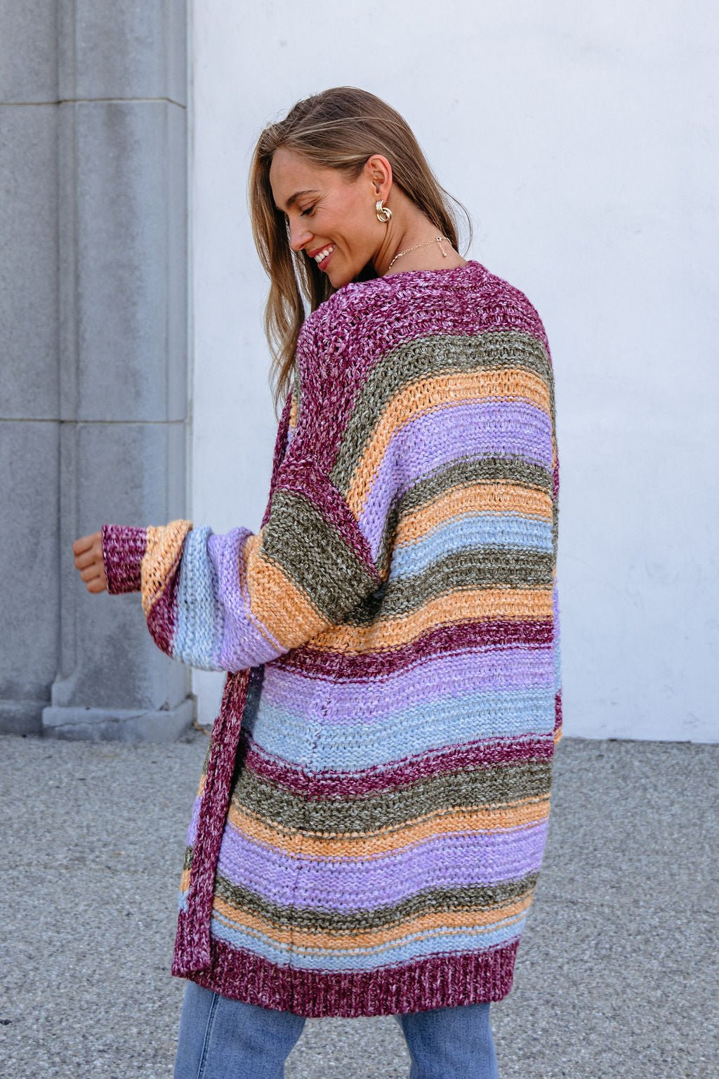 A woman smiles sideways outdoors by a gray column in a Burgundy Striped Open Front Cardigan.