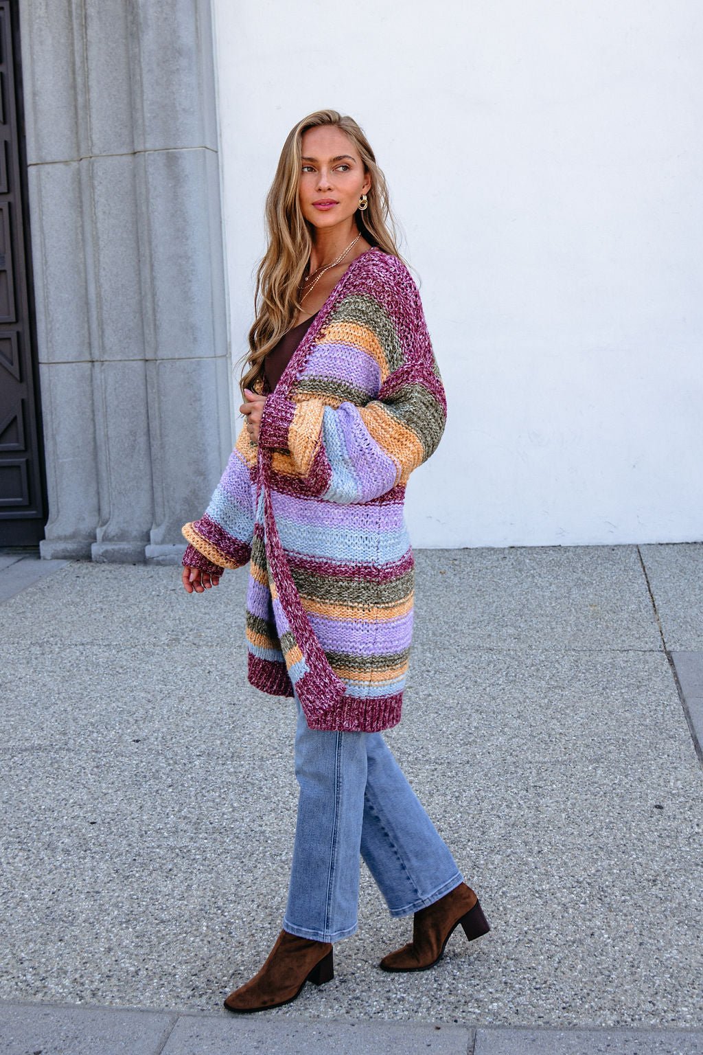 Woman in a Burgundy Striped Open Front Cardigan and blue jeans stands bump-friendly styled by a light wall.