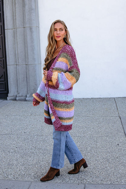 Woman in a Burgundy Striped Open Front Cardigan and blue jeans stands bump-friendly styled by a light wall.