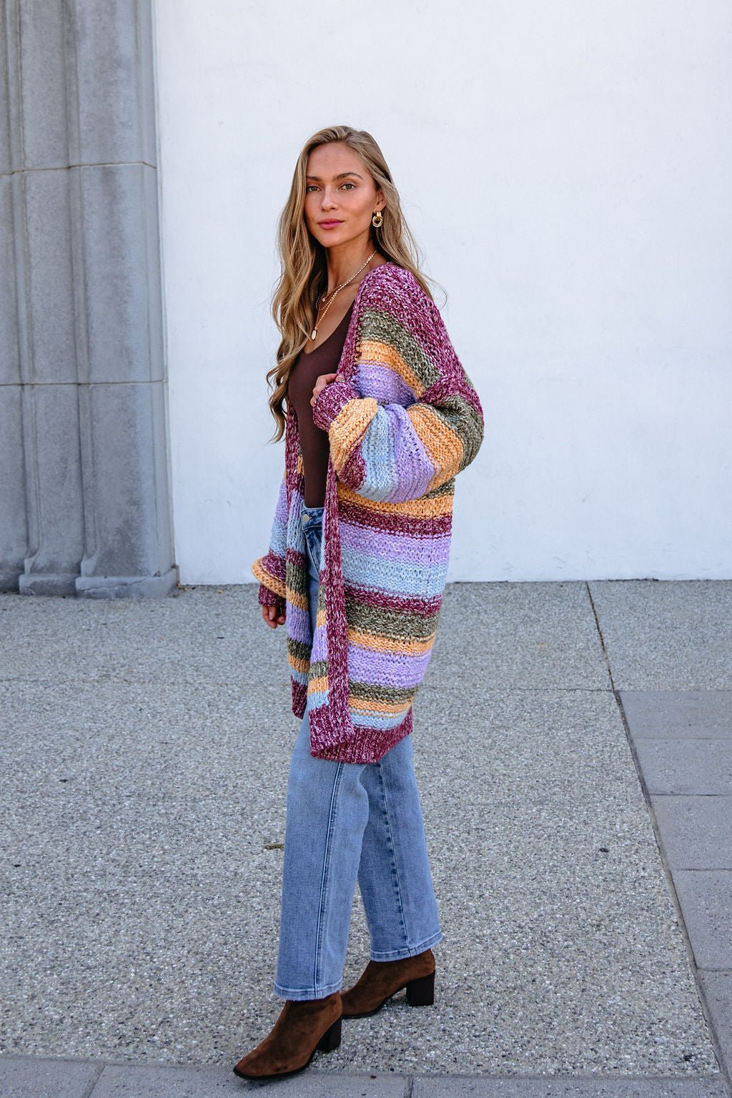 Woman in a Burgundy Striped Open Front Cardigan, brown top, jeans & boots stands outdoors by stone; cardigan is bump-friendly.