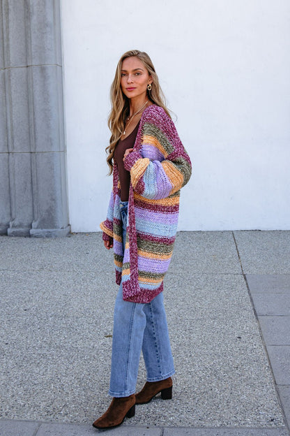 Woman in a Burgundy Striped Open Front Cardigan, brown top, jeans & boots stands outdoors by stone; cardigan is bump-friendly.