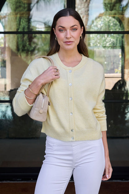 She smiles outside in a Butter Yellow Button Front Cardigan, white pants, and holds a beige handbag.