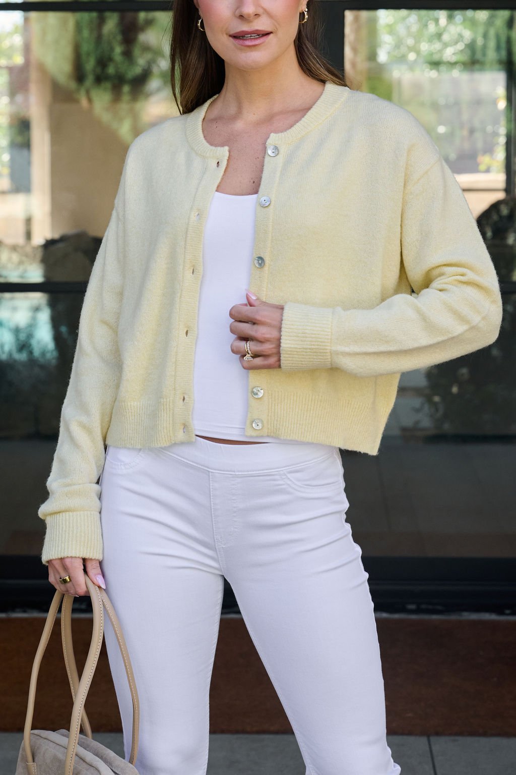 Woman in Butter Yellow Button Front Cardigan over white top and pants, holding beige bag outdoors.