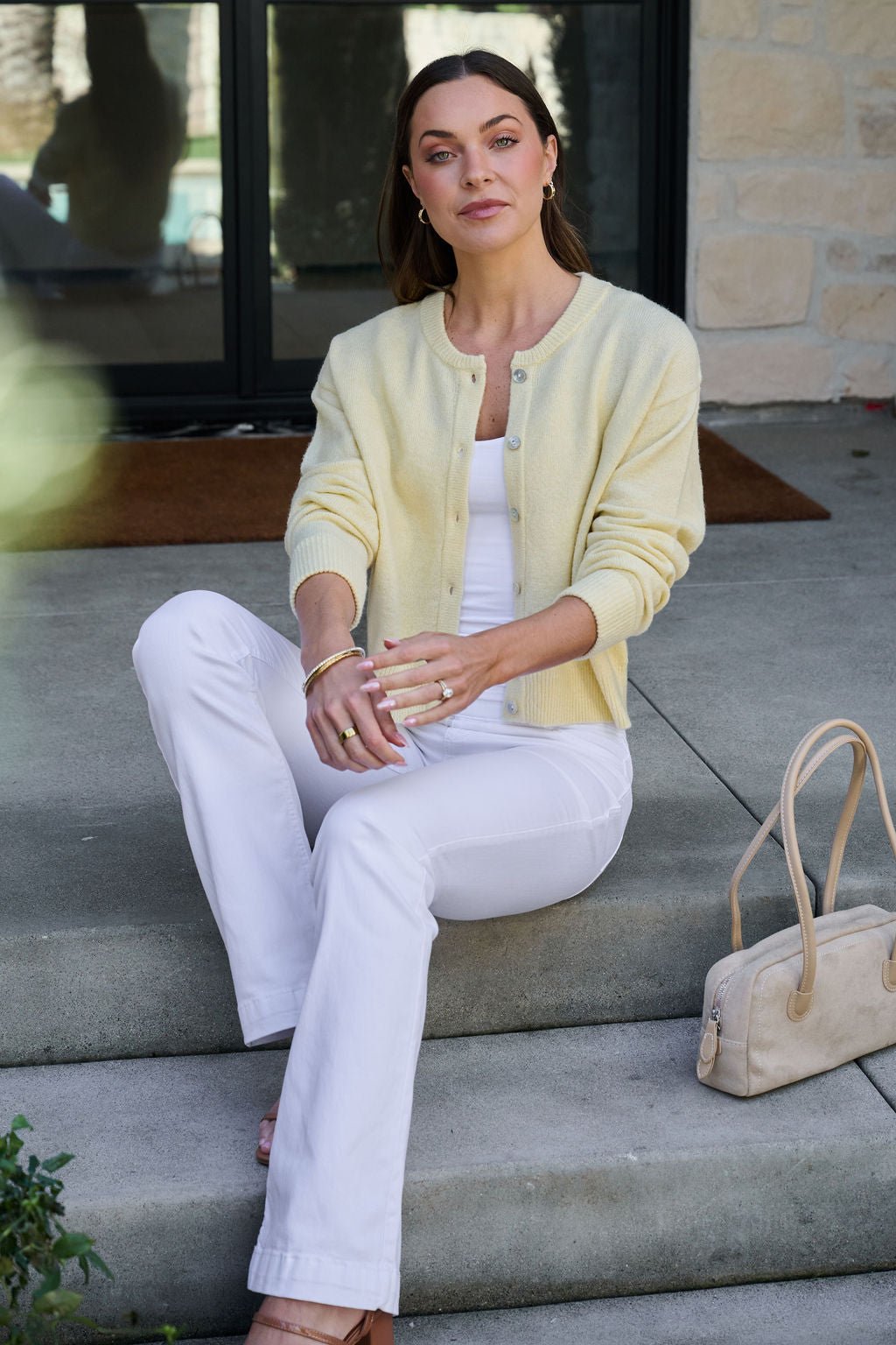 A woman wears the Butter Yellow Button Front Cardigan with white pants, seated on outdoor steps beside a beige handbag.