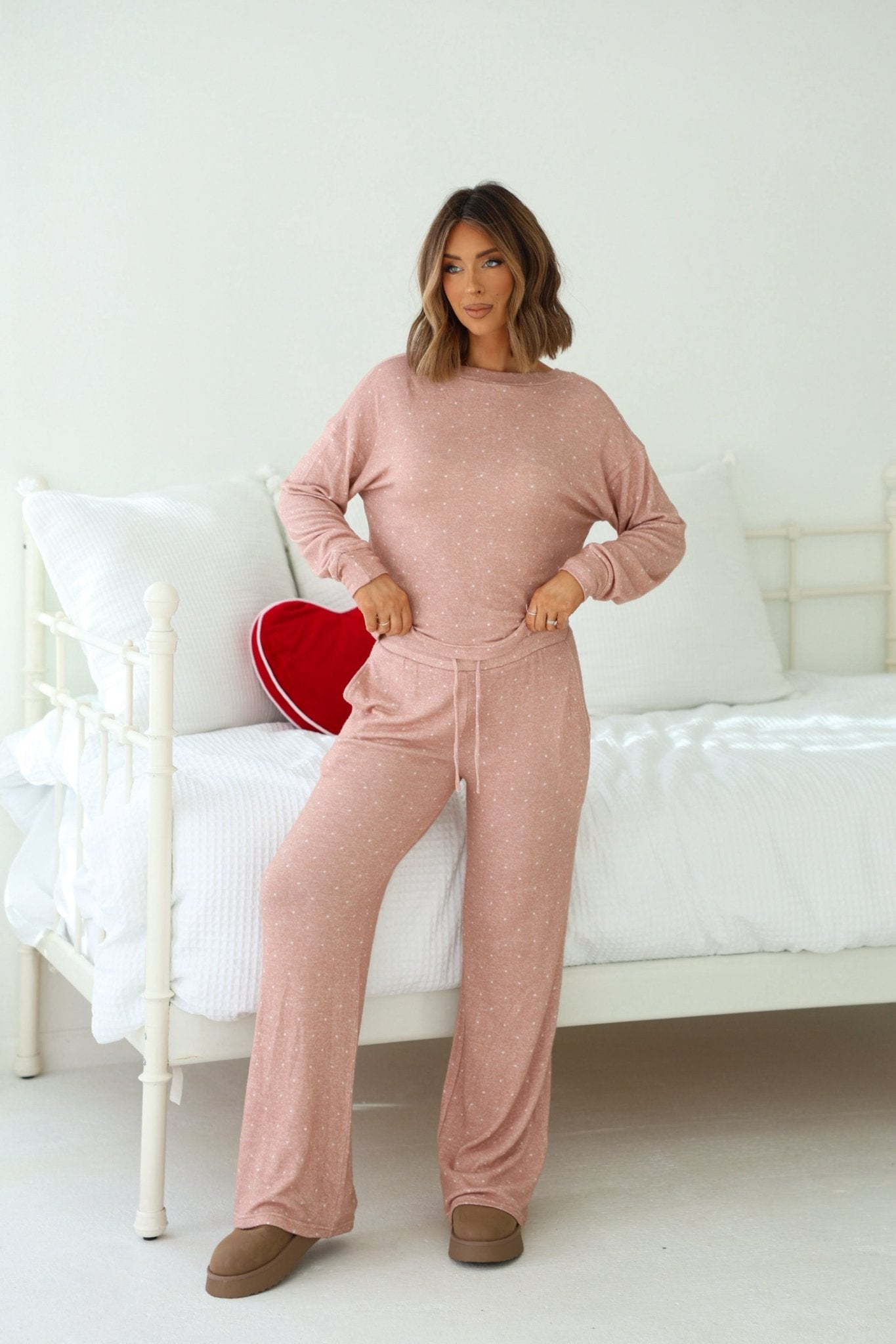 A woman wears the Buttery Soft Blush Polka Dot Lounge Top, standing by a white bed with a red heart pillow in a bright bedroom.