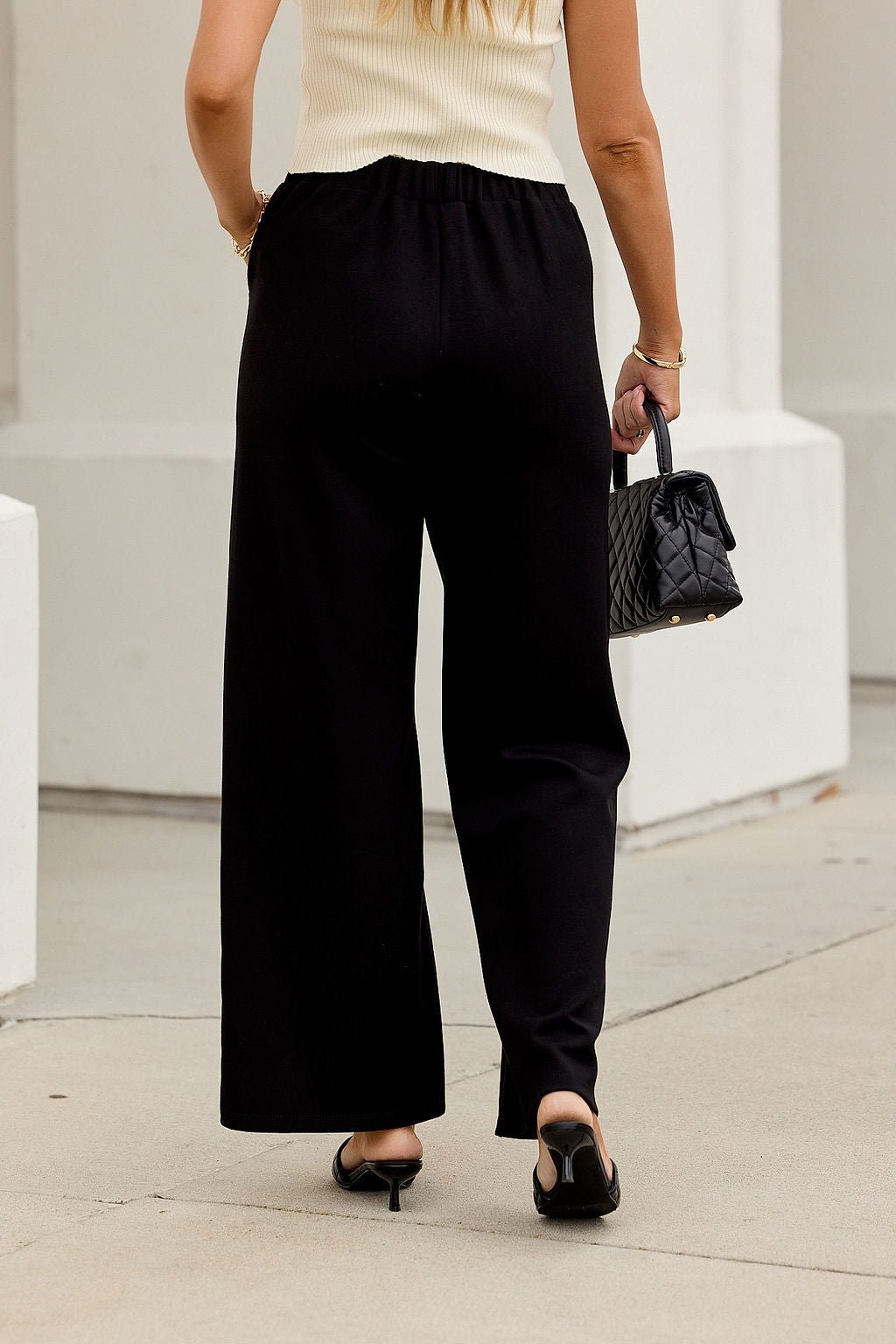 Wearing Buttery Soft Dress Pants - Black, a cream top, and black heels, she carries a black quilted bag on the sidewalk.