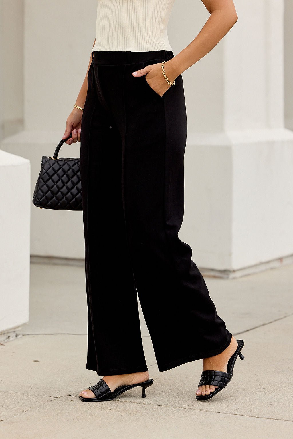 Woman in Buttery Soft Dress Pants - Black, black heeled sandals, cream top, holding a quilted black handbag.