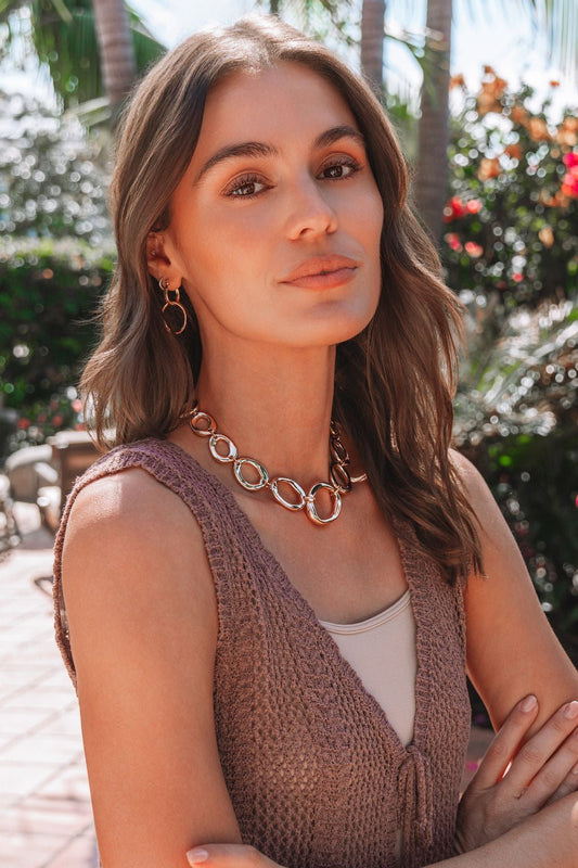 A woman with wavy brown hair wears a Cala Gold Oval Link Necklace and a sleeveless knit top, standing outdoors among greenery.