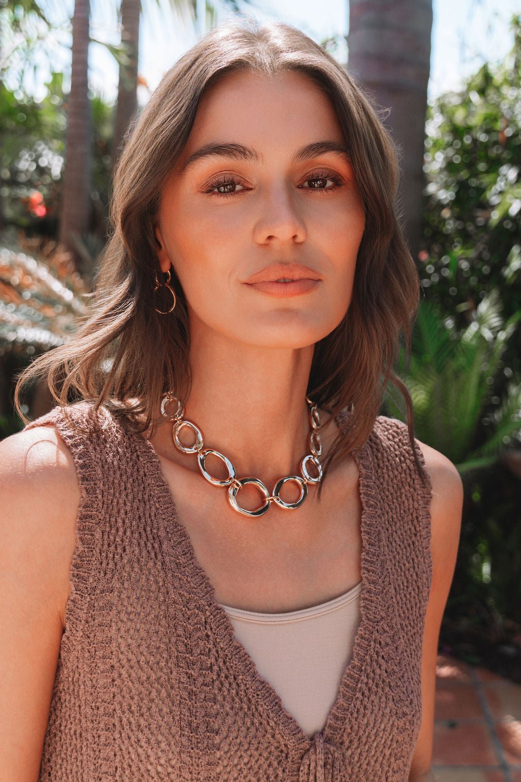 A woman with shoulder-length brown hair wears a Cala Gold Oval Link Necklace and sleeveless knit top, outdoors amid greenery.