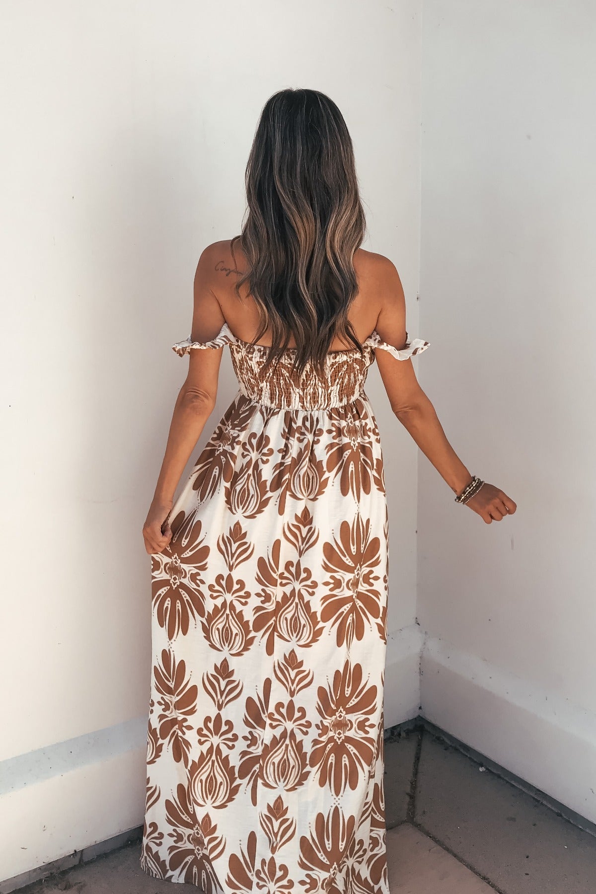Woman with long wavy hair wears the Calista Print Off The Shoulder Maxi Dress - Taupe, standing with her back to the camera.