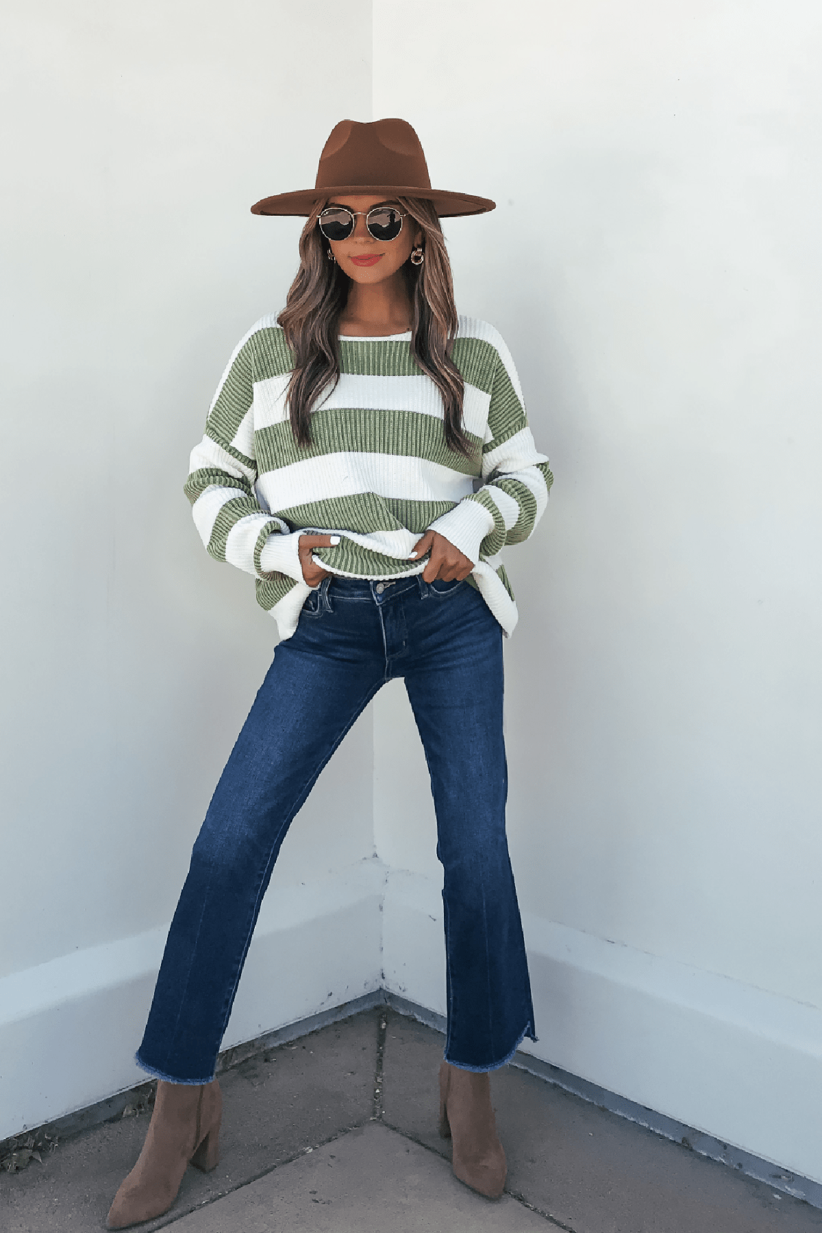 Woman in sunglasses, blue jeans, and tan boots wears a Callahan Green and White Striped Sweater against a white wall.