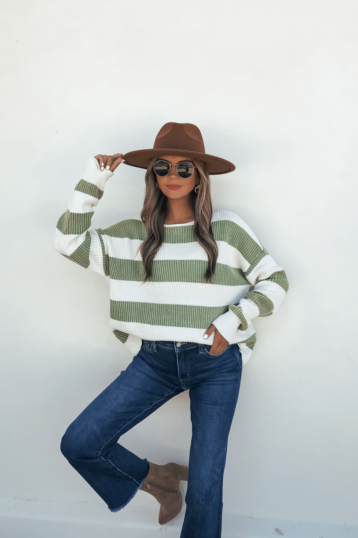 Woman in a wide-brimmed hat, sunglasses, Callahan Green and White Striped Sweater, jeans, and ankle boots poses by a white wall.