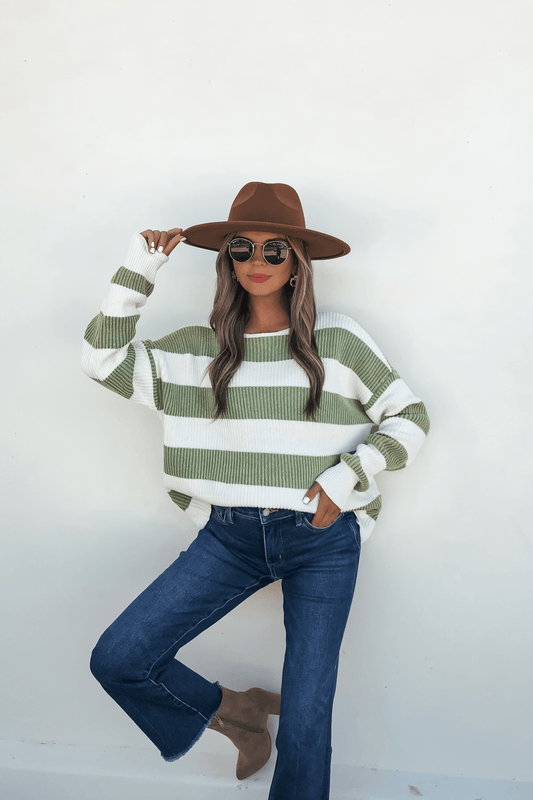 Woman in a wide-brimmed hat, sunglasses, Callahan Green and White Striped Sweater, jeans, and ankle boots poses by a white wall.