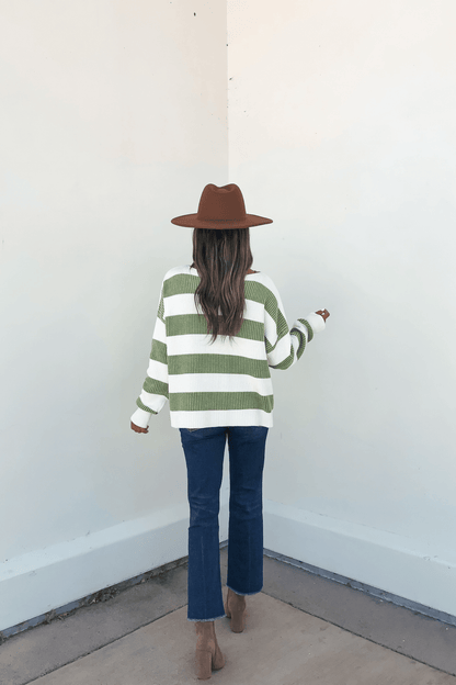 A person in a brown hat, Callahan Green and White Striped Sweater, jeans, and boots stands facing a white wall.