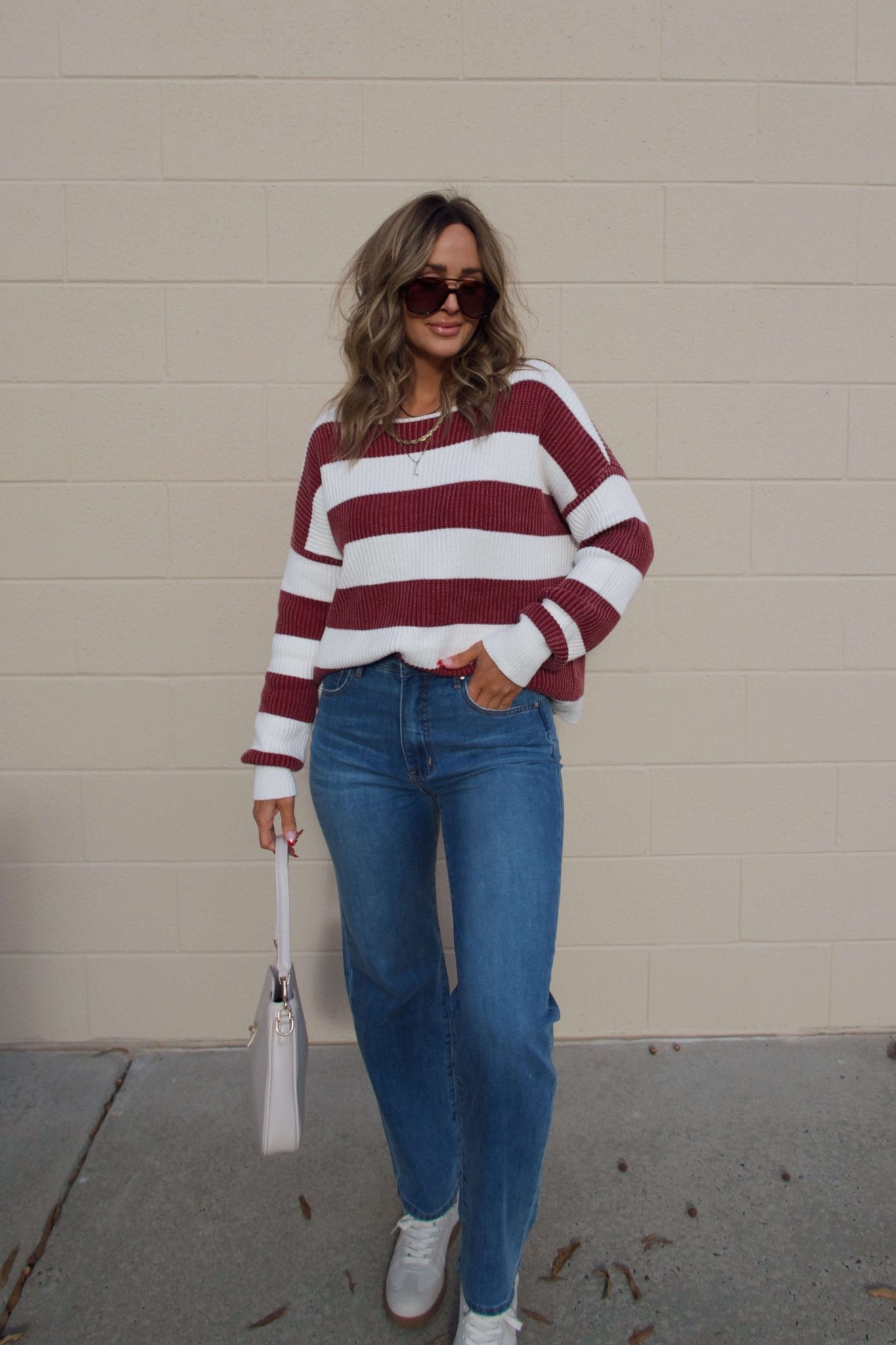 Woman wearing the Callahan Wine and White Striped Sweater, blue jeans, sunglasses, and a light handbag by a beige wall.