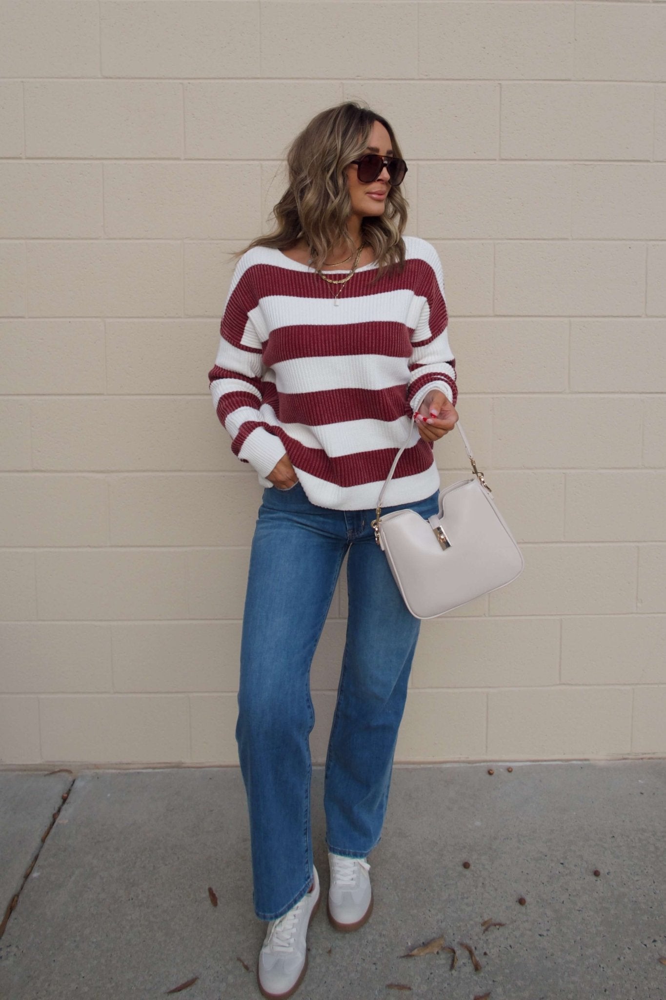 Woman in a cotton knit Callahan Wine and White Striped Sweater, blue jeans, and sunglasses holds a beige handbag by a brick wall.